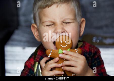 Lustiges Kleinkind leckt weihnachtliche Lebkuchen und macht einen lustigen Gesichtsausdruck aus nächster Nähe. Stimmungsvolles, ästhetisches Zuhause für Silvester. Fröhliches Baby bo Stockfoto
