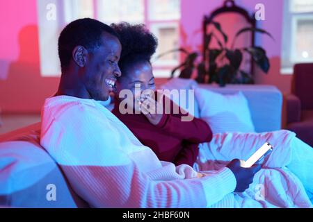 Glückliches afrikanisches Familienpaar lacht, während es sich auf dem Sofa entspannt und auf den Smartphone-Bildschirm schaut Stockfoto