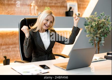Erfolgreiche fröhlich aufgeregt kaukasischen reifen Geschäftsmann, Manager, Aktienhändler, sitzen an einem Tisch im Büro, schreit und freut sich über Erfolg, Sieg oder guten Gewinn, schaut auf einen Laptop, Gesten Hände Stockfoto