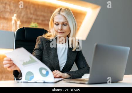 Fokussierte intelligente kaukasische, reife Geschäftsfrau, Finanzanalytikerin, Top-Managerin, sitzt an ihrem Schreibtisch im Büro, studiert Finanzdiagramme, analysiert und plant Finanzstrategie Stockfoto