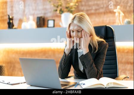 Müde verärgert kaukasisch reife blonde Frau, ceo oder Manager, in Anzug, müde von Online-Arbeit auf Laptop, erleben Stress und Kopfschmerzen, Pause von der Arbeit, massieren Tempel, Ruhe brauchen, Augen geschlossen Stockfoto