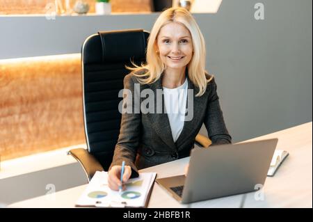 Das Porträt einer ziemlich reifen Mitarbeiterin sitzt am Schreibtisch im Büro und arbeitet mit Dokumenten und einem Laptop. Eine elegante Geschäftsfrau mittleren Alters in stilvollem Anzug blickt freundlich lächelnd auf die Kamera Stockfoto