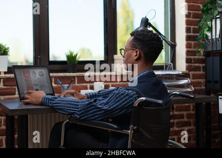 afroamerikanischer Mitarbeiter, der gelähmt ist und Leistungsindikatoren auf einem Laptop in einem Red Brick Office analysiert. Startup-Besitzer mit Blick auf tragbaren Computer mit Verkaufsstatistiken. Stockfoto