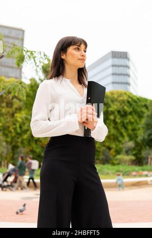 Outdoor-Foto von jungen attraktiven zuversichtlich europäischen Geschäftsfrau in formellen Outfit mit Zwischenablage wartet auf Client bei der Sitzung, erfolgreiche kaukasische Geschäftsfrau auf der Straße der Stadt stehen Stockfoto