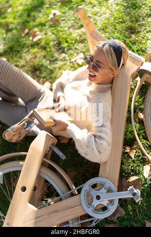 Von oben der Frau in der Sonnenbrille lachend mit geöffnetem Mund, während sie auf Gras lag und sich auf dem Holzfahrrad im Park lehnte Stockfoto