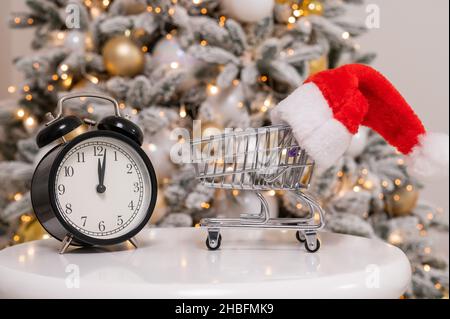 Weihnachtsgeschenke Zeit zum Einkaufen. Ein Einkaufswagen mit Weihnachtsmütze und Wecker am Weihnachtsbaum. Stockfoto