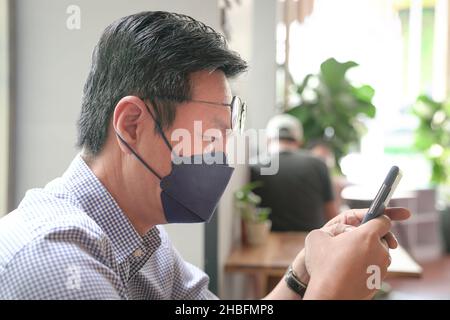 Asiatischer Mann mit Gesichtsmaske, der sein Telefon benutzt, SMS in einem Restaurant. Stockfoto