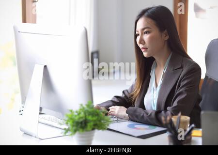 Attraktive asiatische Geschäftsfrau, die sich auf ihr Marketingprojekt auf den pc an ihrem Schreibtisch konzentriert. Stockfoto