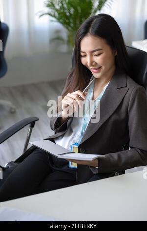 Porträt einer asiatischen jungen Chefin, die im modernen Büro sitzt und auf ihrem Notizbuch liest. Prüfen, erneut prüfen, Terminplanbuch prüfen. Stockfoto