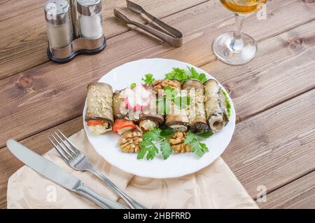 Gesunde Auberginen-Brötchen gefüllt mit Käse, serviert mit Gemüse und Kräutern auf einem weißen Teller auf dunklem Holzboden Stockfoto