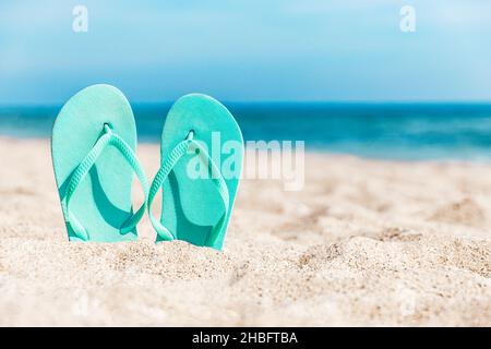 Türkisfarbene oder hellblaue Flip Flops oder Sandalschuhe stecken im Sand am Sandstrand an der Küste oder am Meer. Reise- oder Urlaubskonzept in heißen Ländern im Sommer oder Winter. Hochwertige Fotos Stockfoto