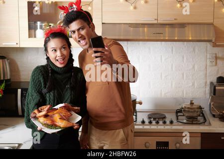 Aufgeregt fröhlich junges Paar Video ruft Freunde oder Familienmitglieder und zeigt gegrilltes Huhn zum Weihnachtsessen Stockfoto
