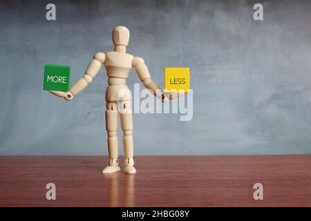 Hölzerne menschliche Figur balanciert Holzwürfel mit Text MEHR und WENIGER. Stockfoto