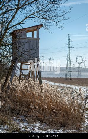 Holzjagd Turm in der Nähe des landwirtschaftlichen Feldes im Winter. Huntsman Hochsitz mit Hochspannungsleitungen und Strommasten. Stockfoto