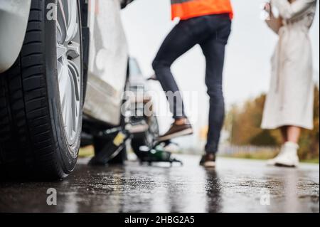 Nahaufnahme eines Autos auf verregneter Straße mit einer Fahrerin und einer Assistenzarbeiterin vor unscharfem Hintergrund. Männliche Auto Mechaniker Reparatur Frau Auto auf der Straße. Fokus auf Care Wheel. Stockfoto