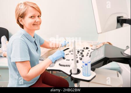Sonograf, der weibliche Patienten mit einem Ultraschallscanner untersucht. Ärztin in blauem Hemd, die Kamera anschaut und lächelt, während sie Ultraschalluntersuchung macht. Konzept der Gesundheitsfürsorge und Ultraschalldiagnostik. Stockfoto