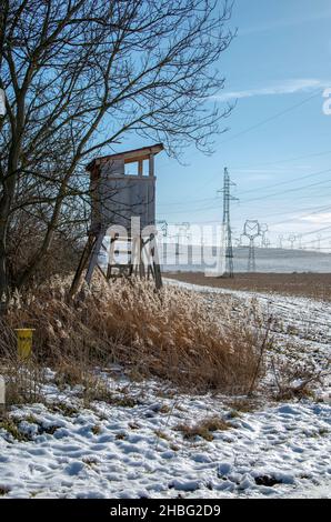 Holzjagd Turm in der Nähe des landwirtschaftlichen Feldes im Winter. Huntsman Hochsitz mit Hochspannungsleitungen und Strommasten. Stockfoto