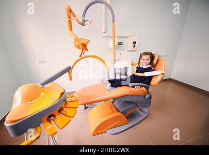 Nettes weibliches Kind sitzt in gelben Zahnarztstuhl. Liebenswert Kind in Schuhüberzügen warten auf Verfahren in der Zahnarztpraxis. Konzept der Kinderstomatologie, Zahnmedizin und Zahnpflege. Stockfoto