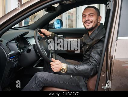 Ein hübscher junger Mann, der sich im Autohaus für ein neues Auto entschieden hat, in einem Auto sitzt und den Daumen nach oben zeigt. Typ in legerer Kleidung und goldener Uhr. Stockfoto
