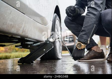 Nahaufnahme der männlichen Hände, die das Fahrzeug mit dem Wagenheber anheben, bevor der Reifenpanne gewechselt wird. Junger Mann mit Uhr am Handgelenk mit speziellen Gerät für das Heben Auto, während die Reparatur Frau Fahrzeug auf der Straße. Stockfoto