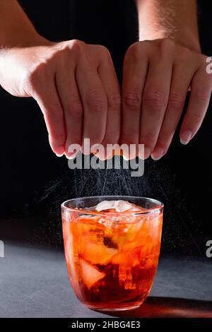 Barkeeperin drückt Öle aus einer Orangenschale für einen Cocktail auf dunklem Hintergrund aus. Nahaufnahme von Händen und Spritzern Stockfoto