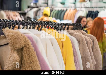 Mode Kunstpelzmäntel hängen auf Rack im Bekleidungsgeschäft, verschiedene Farben, selektive Fokus, verschwommener Hintergrund des Ladeninneren, Mädchen, Kunden Stockfoto