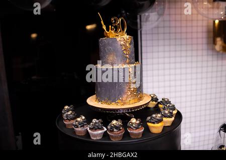 Festlicher zweistufiger schwarzer Kuchen mit Golddeko zum 30th-jährigen Jubiläum. Viele Cupcakes aus Schokolade und Vanille mit luxuriöser Dekoration. Candybar für BI Stockfoto