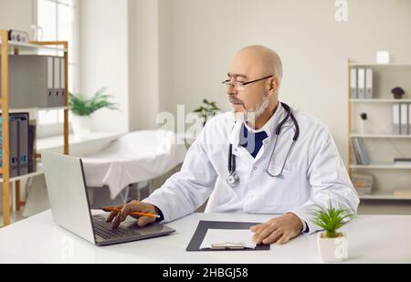 Leitender Arzt in Brille und weißem medizinischen Kleid, der am Laptop am Arbeitsplatz im Büro sitzt. Stockfoto