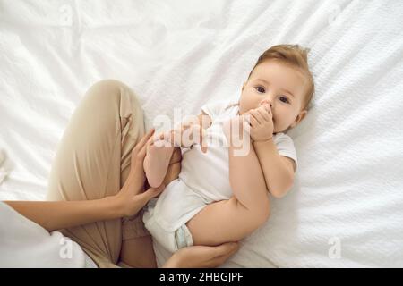 Porträt von niedlichen charmanten sechs Monate alten Mädchen spielen mit ihren Beinen auf dem Rücken auf weißem Bett liegen. Stockfoto