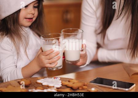 Portrait von schönen Baumjahr Mädchen mit ihrer Mutter in santa Hut sitzen am Tisch vor der Küche geschmückt Lichterketten und trinken frische Milch Stockfoto