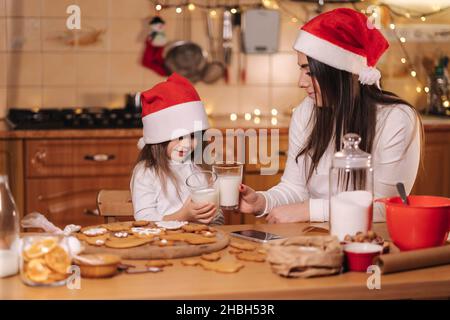 Portrait von schönen Baumjahr Mädchen mit ihrer Mutter in santa Hut sitzen am Tisch vor der Küche geschmückt Lichterketten und trinken frische Milch Stockfoto