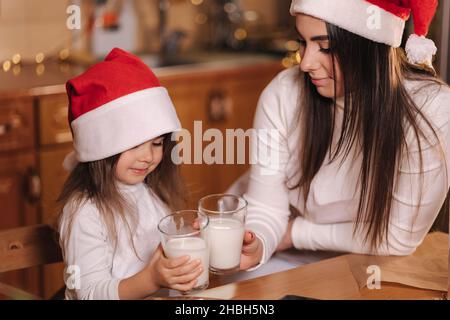 Portrait von schönen Baumjahr Mädchen mit ihrer Mutter in santa Hut sitzen am Tisch vor der Küche geschmückt Lichterketten und trinken frische Milch Stockfoto