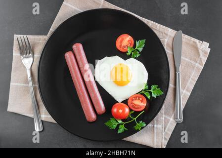 Herzförmige Rühreier mit Würstchen auf schwarzem Teller zum Valentinstag. Stockfoto