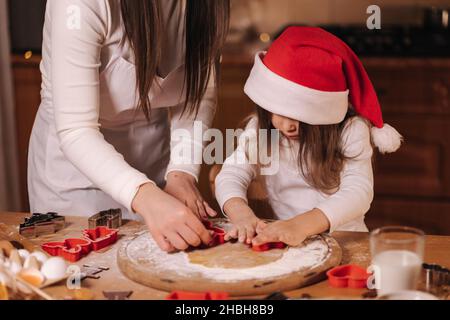 Lebkuchen zu Hause machen. Kleines Mädchen mit ihrer Mutter, die Kekse aus Lebkuchenteig schneidet. Weihnachts- und Neujahrstraditionen Konzept. Weihnachtsbäckerei Stockfoto