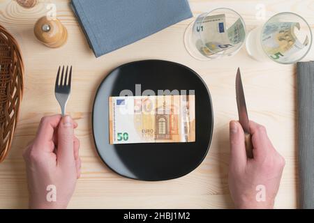 Konzeptuelle Studioaufnahme des Esstisches mit Euro-Banknoten auf dem Teller statt des Essens. Konzept für steigende Lebensmittelpreise, Inflation, Wirtschaftskrise Stockfoto