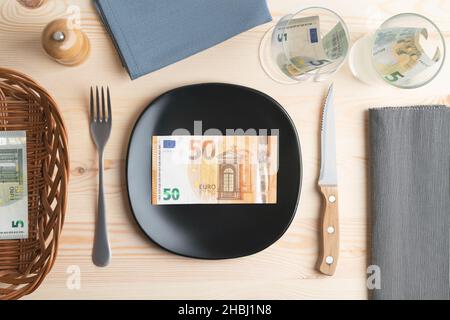 Konzeptuelle Studioaufnahme des Esstisches mit Euro-Banknoten auf dem Teller statt des Essens. Konzept für steigende Lebensmittelpreise, Inflation, Wirtschaftskrise Stockfoto