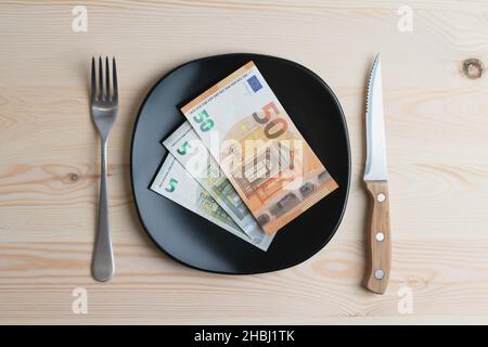 Konzeptuelle Studioaufnahme des Esstisches mit Euro-Banknoten auf dem Teller statt des Essens. Konzept für steigende Lebensmittelpreise, Inflation, Wirtschaftskrise Stockfoto
