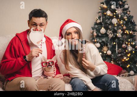 Porträt einer glücklichen Familie mit einem Welpen, der festliche Party-Requisiten für einen Fotostand hält. Paar Frau und Mann in Santa Hut und Hund in Pullover Spaß auf Stockfoto