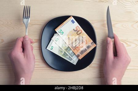 Konzeptuelle Studioaufnahme des Esstisches mit Euro-Banknoten auf dem Teller statt des Essens. Konzept für steigende Lebensmittelpreise, Inflation, Wirtschaftskrise Stockfoto