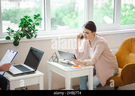 Geschäftspartnerinnen arbeiten von zu Hause aus mit einem Laptop, während sie im Sessel sitzen. Frau, die am Telefon spricht. Fernarbeiten. Stockfoto