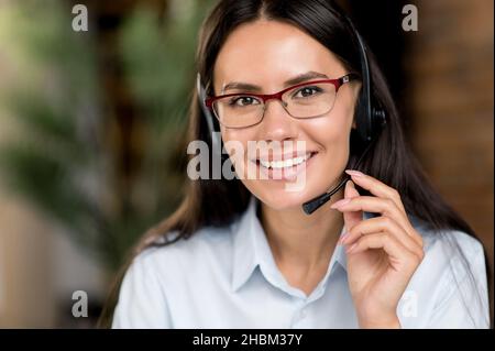 Nahaufnahme Portrait nette fröhliche kaukasische Brünette Geschäftsfrau, Berater oder Top-Manager, erfolgreiche Immobilienmakler, trägt Brille und Headset, in formeller Kleidung, Blick auf die Kamera, lächelnd Stockfoto