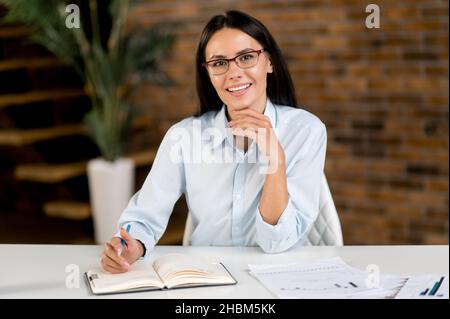 Schöne intelligente erfolgreiche junge Erwachsene Brünette kaukasischen Geschäftsfrau, ceo oder Broker trägt Brille, sitzen am Schreibtisch im modernen Büro, Blick auf die Kamera, freundlich lächelnd Stockfoto