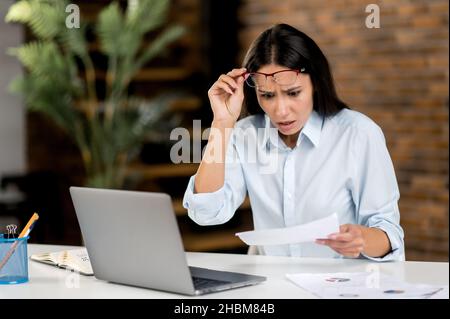 Schockiert aufgeregte junge Frau, CEO, Manager oder Makler mit Brille sitzt an einem Tisch in einem modernen Büro, schaut sich überraschend Dokumente an, schockiert von Statistiken und Verlusten, finanziell ruiniert Stockfoto