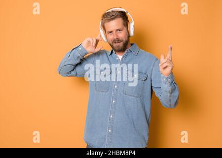 Junger Mann, der Kopfhörer trägt und Musik hört, steht isoliert auf orangefarbenem Hintergrund. Stockfoto