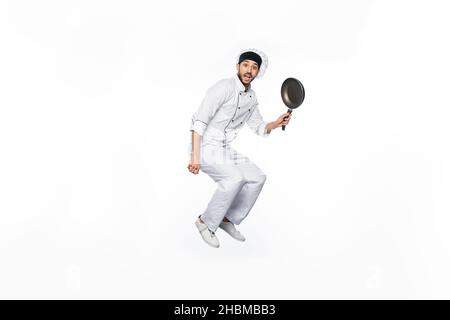 Begeisterter Koch in Hut und Uniform springen und halten Bratpfanne isoliert auf weiß Stockfoto