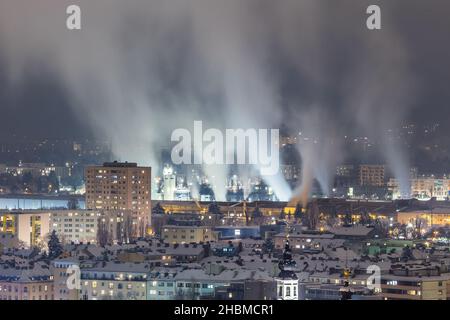 Nächtlicher Blick auf die Stadt mit Gebäuden und einem rauchenden Stahlwerk mit beeindruckenden Abgasen Stockfoto