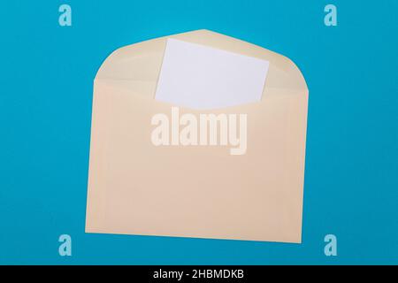 Beigefarbener Umschlag mit leerem weißen Blatt Papier im Inneren, auf blauem Hintergrund liegend - Mock Up with Copy Space. Posteingang oder Benachrichtigung, Postkarte senden - Draufsicht, Flat Lay Stockfoto