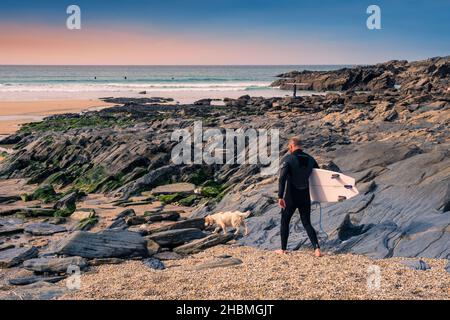 Ein Surfer, der sein Surfbrett trägt und mit seinem Hund über Felsen zum Meer bei Little Fistral in Newquay in Cornwall läuft. Stockfoto