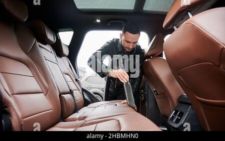 Blick von innen auf einen jungen Mann, der im Freien an der Autowaschanlage Wasch fährt. Hübscher Fahrer, der den Innenraum des Fahrzeugs mit einem Staubsauger reinigt. Stockfoto