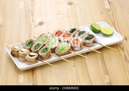 Japanisches Essen. Gemüse, das in dünn geschnittenes Schweinefleisch eingewickelt ist, wird Spieße und gegrillt. Junger Mais, Knoblauchsprossen, Salat, Tomaten und Koriander. Stockfoto